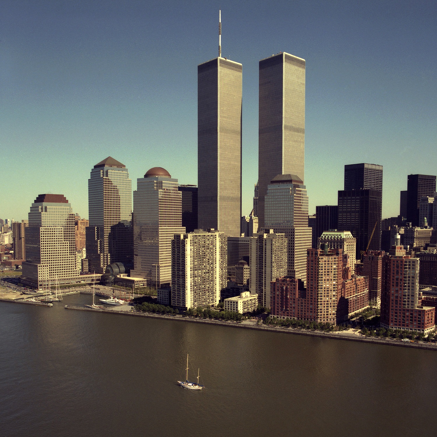A historical aerial photo of lower Manhattan shows the Twin Towers rising above the World Trade Center site on a sunny day. A sailboat passes by on the Hudson River in the foreground.