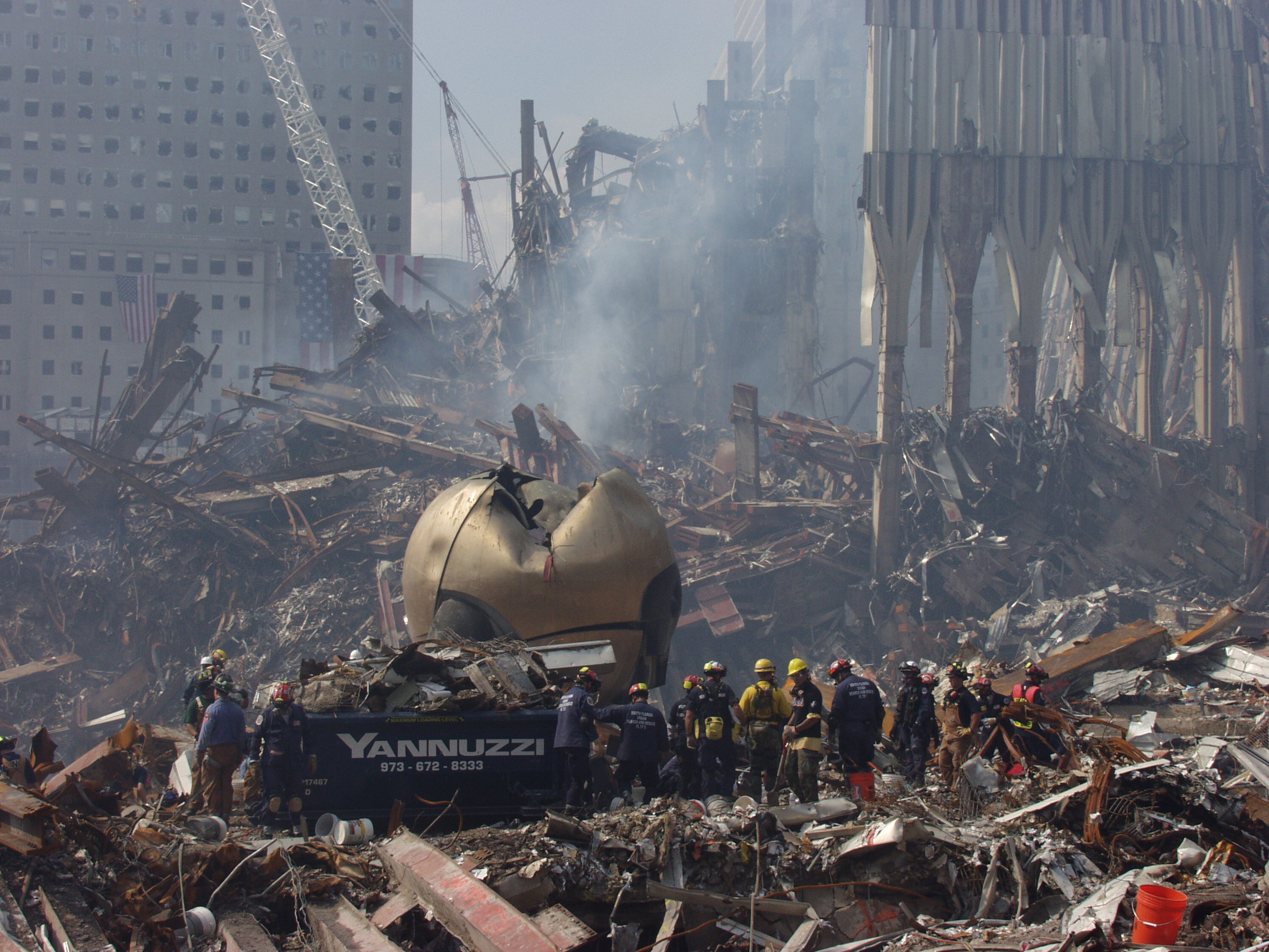 The heavily damaged Koenig Sphere stands in the debris of Ground Zero after the 9/11 attacks. Dozens of rescue workers stand around the sphere. The rubble of the Twin Towers can be seen in the background.