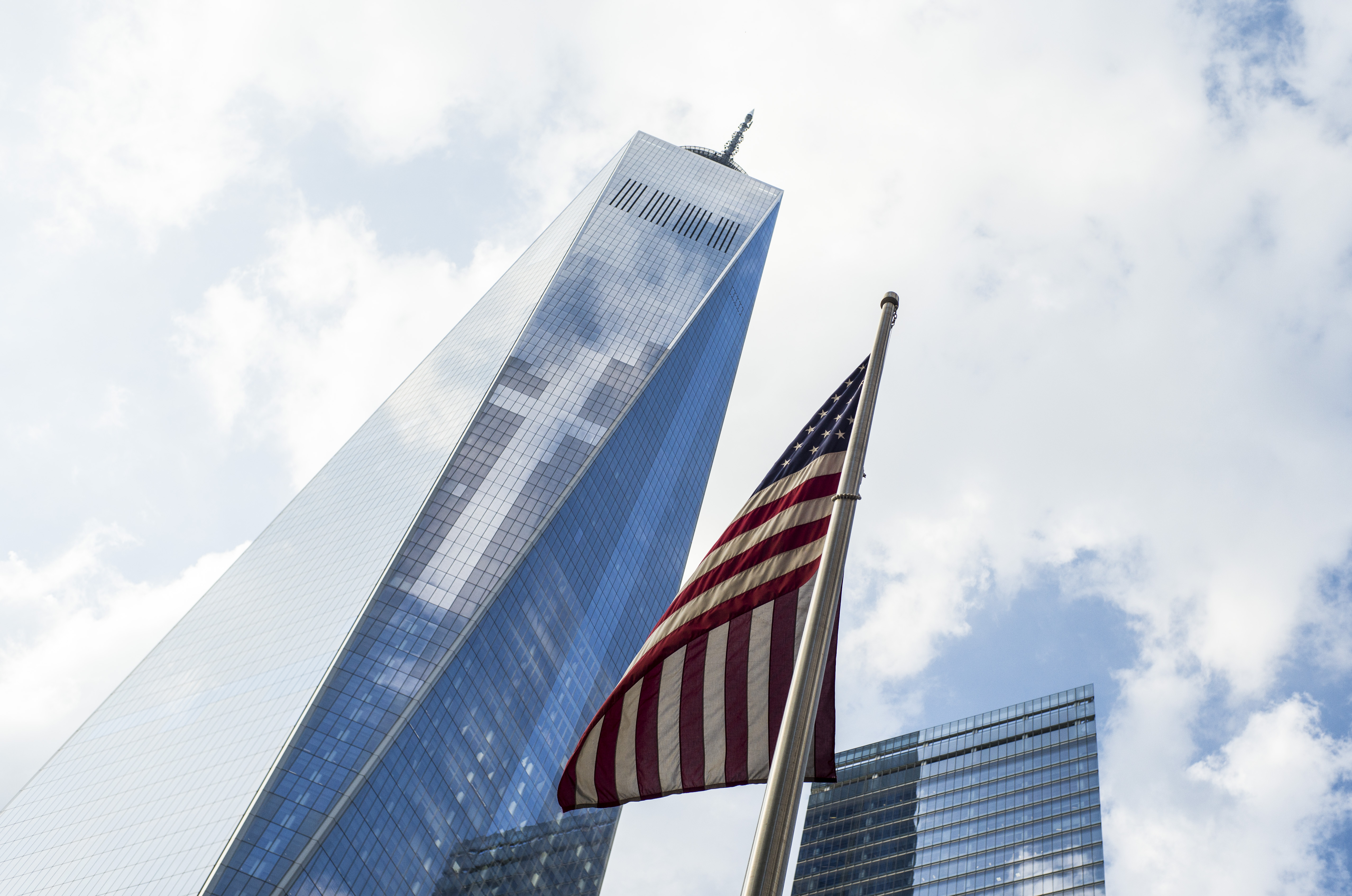 An American flag flies at the 9/11 Memorial as One World Trade Center towers before a partly cloudy sky in the background.