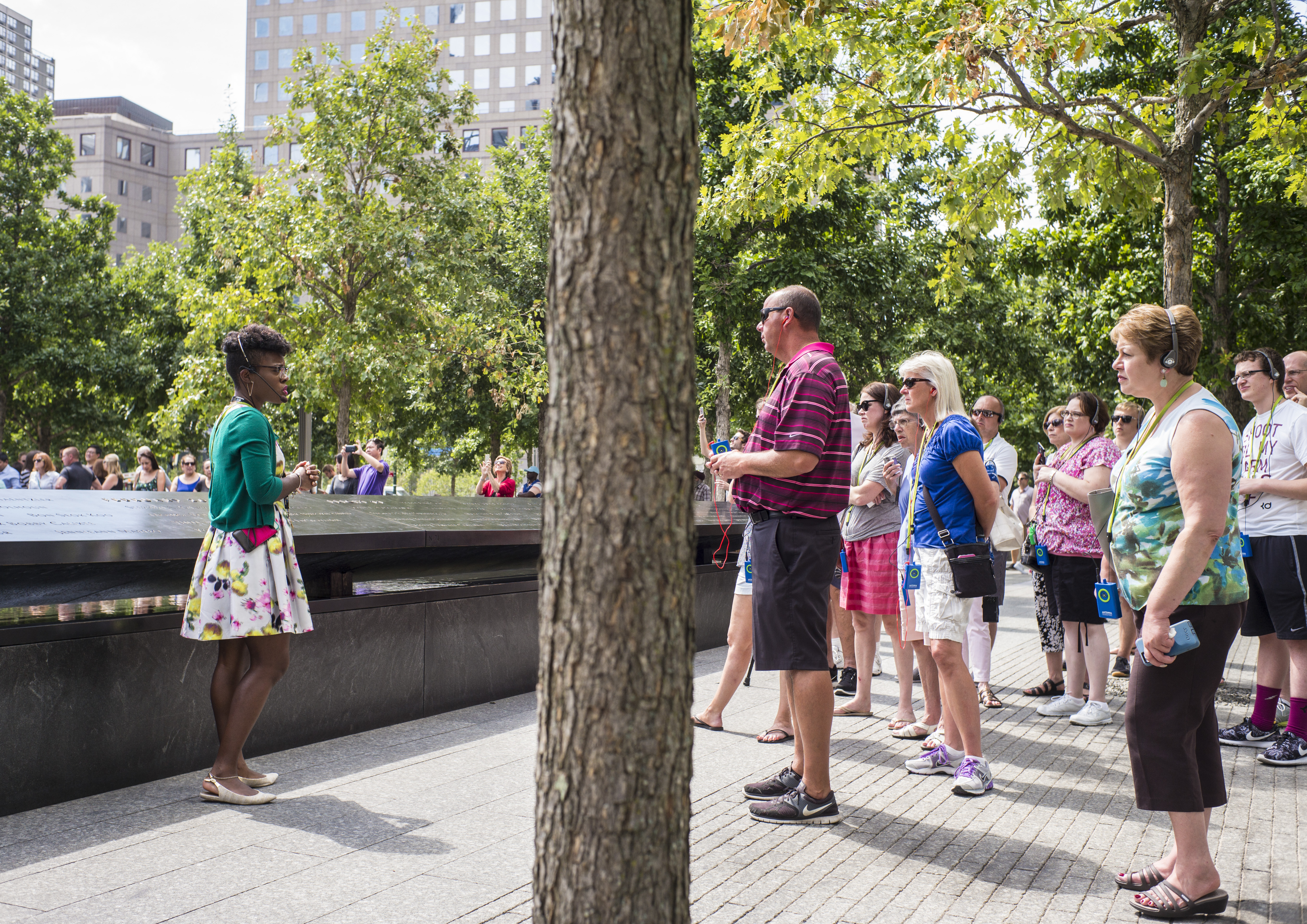 Nicole Richardson leads a tour on the 9/11 Memorial plaza. A group of visitors stand and watch her as she speaks in front of a reflecting pool on a sunny day.
