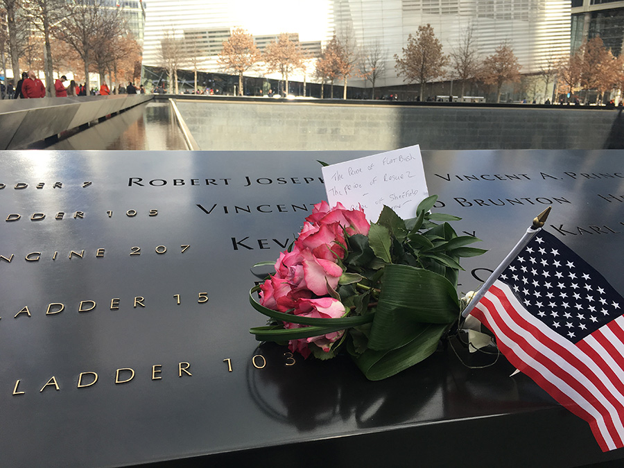 A bundle of flowers, a note, and a small American flag have been placed at a name on a bronze parapet at the south pool of the Memorial. The Museum pavilion can be seen in the background along with trees on the plaza.