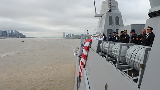 Members of the Navy and other people in official uniforms are seen on the USS New York as the ship travels down the Hudson River in this view looking south. The skylines of lower Manhattan and Jersey City are visible in the distance on a cloudy day.