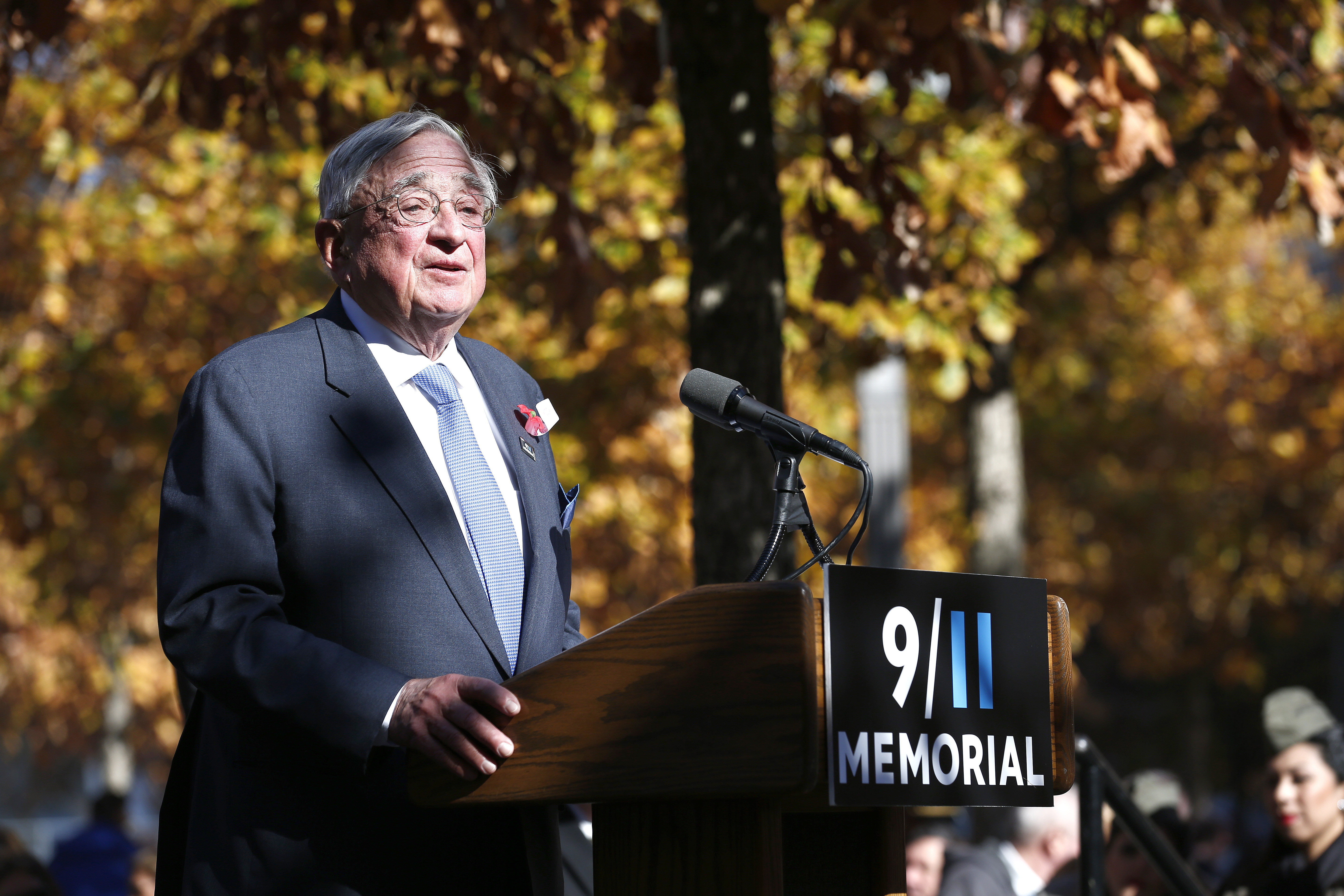 John E. Zuccotti wears a suit and tie as he speaks at a podium on the 9/11 Memorial.