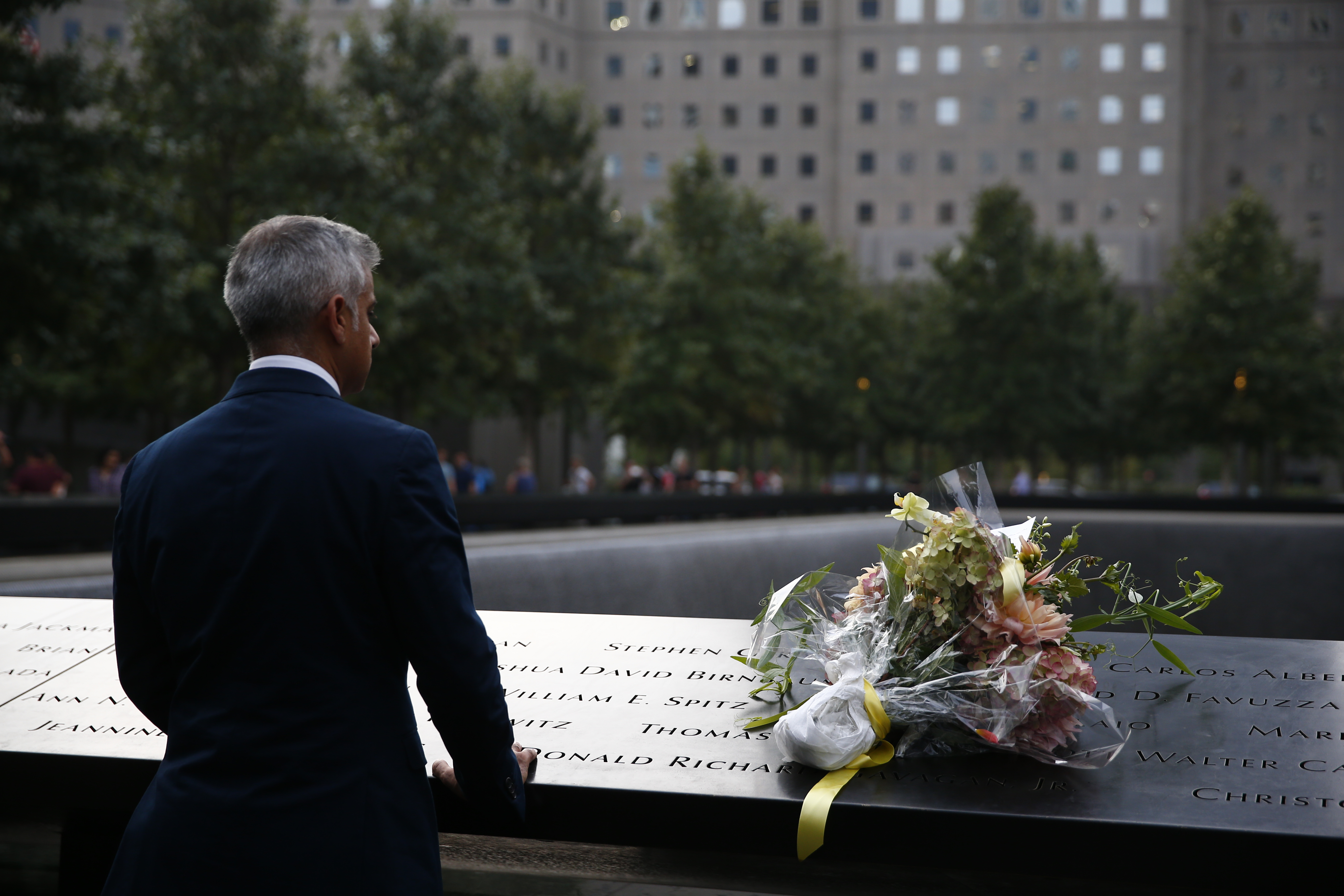 Shadiq Khan, the mayor of London, looks out at a reflecting pool at the 9/11 Memorial. A bouquet of flowers sits on the bronza parapet beside him.