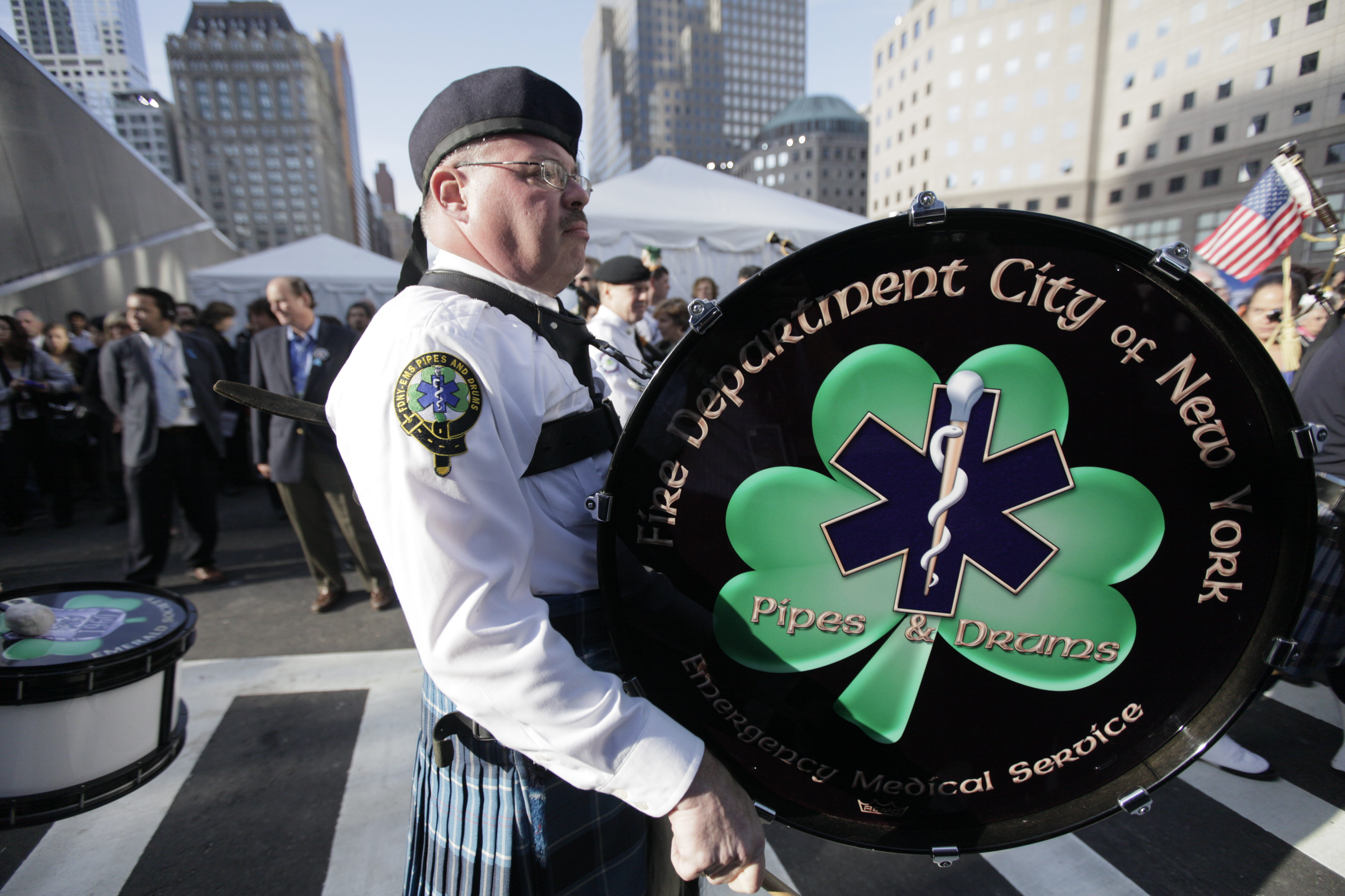 Members of the FDNY Emerald Society Pipes and Drums band play at the 9/11 Memorial during an event. White tents and groups of people are visible in the background.