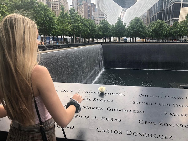Caleigh Leiken’s back is turned to the camera as she places a white rose at the name of Alena Sesinova at the 9/11 Memorial. Water cascades down the sides of a reflecting pool in the background.