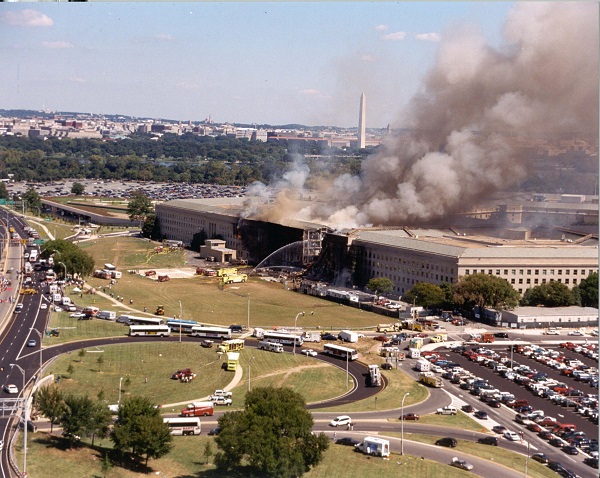 Thick smoke rises from the Pentagon on 9/11 as emergency responders converge on the scene. A charred hole can be seen where American Airlines Flight 77 was flown into the building. The skyline of Washington, D.C. is visible in the background.