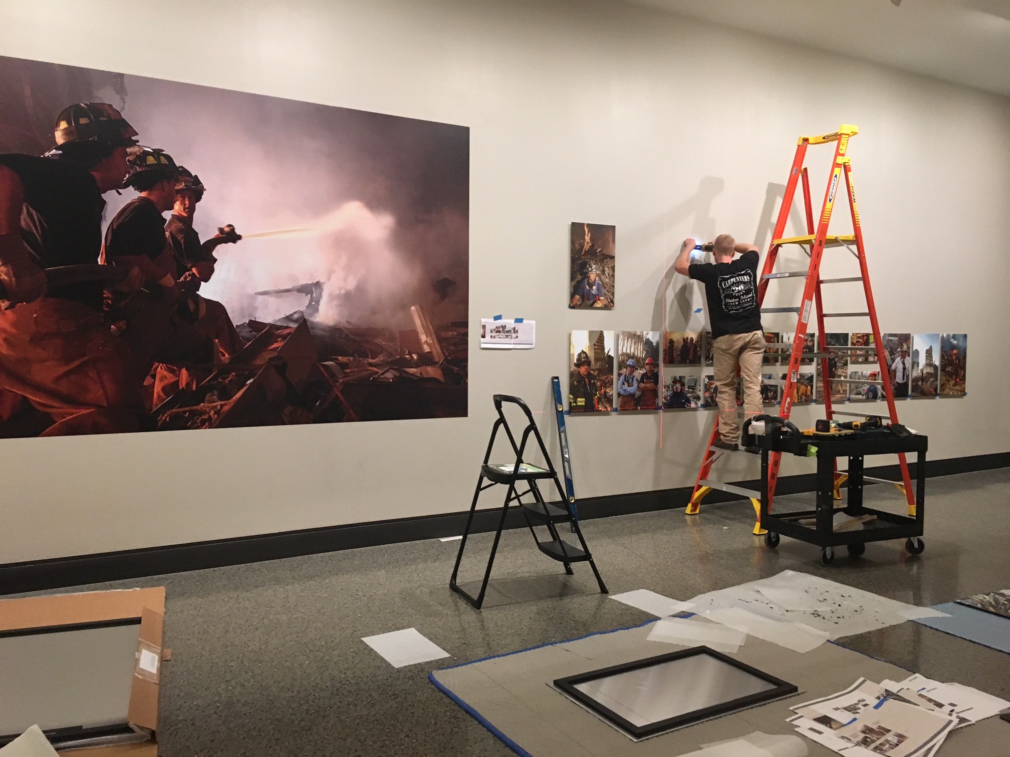 A man stands on an orange ladder as he installs the exhibition “Hope at Ground Zero.” About a dozen FEMA photographs of Ground Zero have been positioned on a white wall in front of the man.