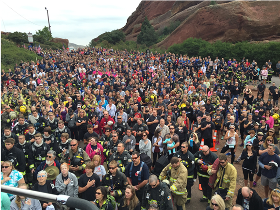 Hundreds of people put their right hands on their heart in a view over the Colorado Memorial Stair Climb on a cloudy day. Many first responders in the crowd are dressed in bunker gear and other uniforms.