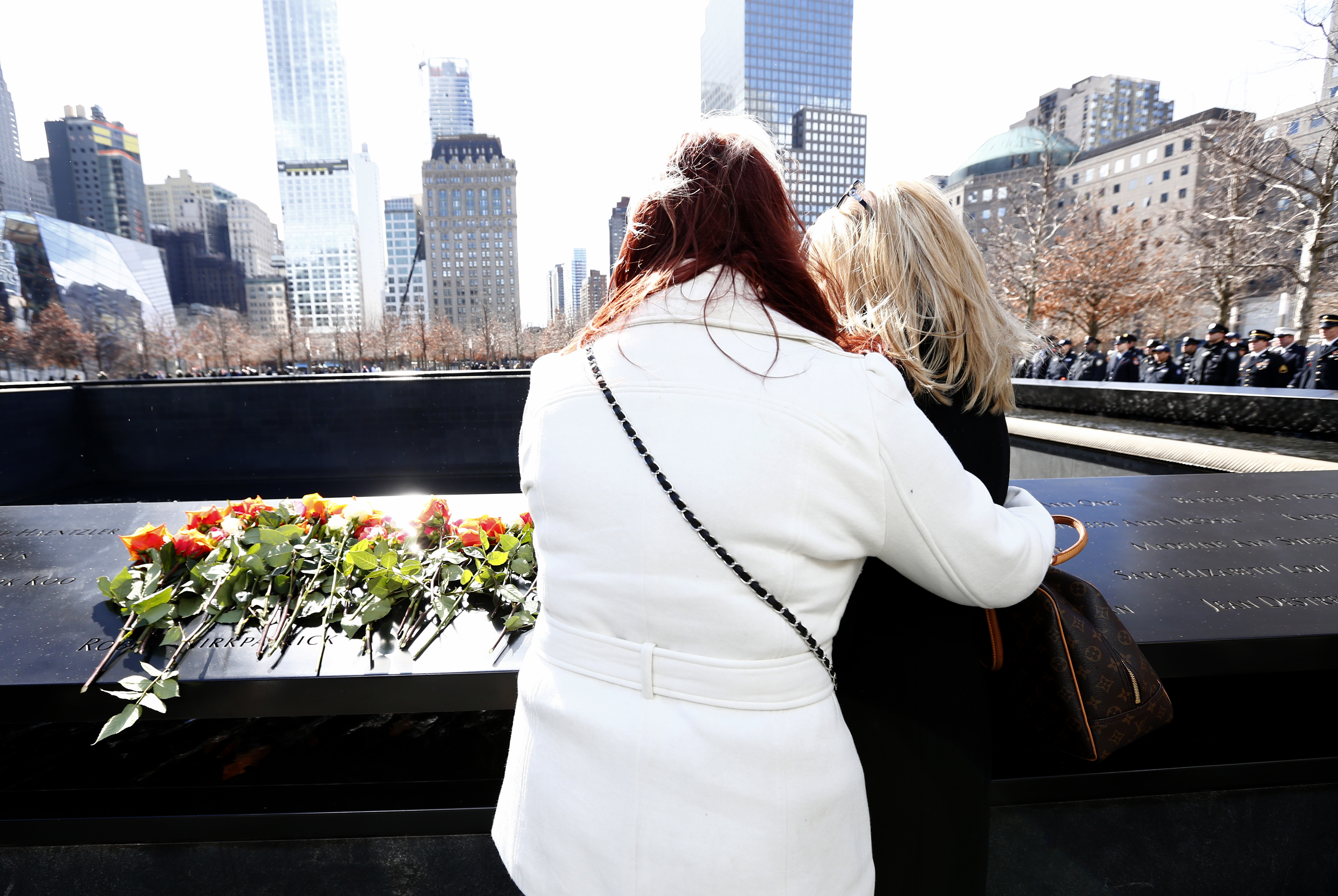 Two women, one in a white jacket and the other in a black jacket, embrace as they view a name at the Memorial. Flowers have been placed on the bronze parapet where the names are inscribed. 