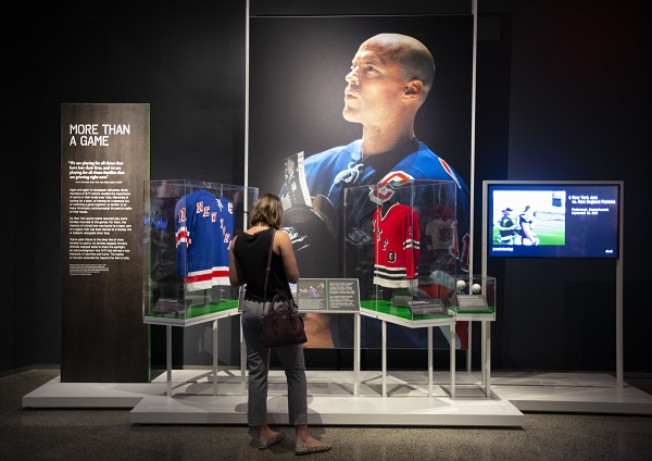 A woman stands in front of a large photo of Rangers captain Mark Messier at the exhibition, Comeback Season: Sports After 9/11. Jerseys and other sports artifacts are displayed in front of the photo of Messier.