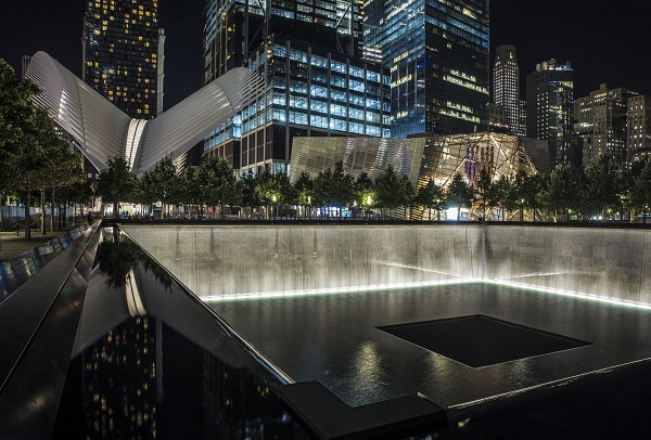The North Tower reflecting pool is seen illuminated at night. The Oculus transportation hub and the Museum pavilion are seen in the background, along with other buildings of the lower Manhattan skyline.