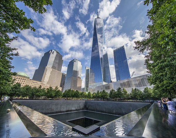 One World Trade Center towers over the south pool of Memorial plaza on a partly cloudy day.