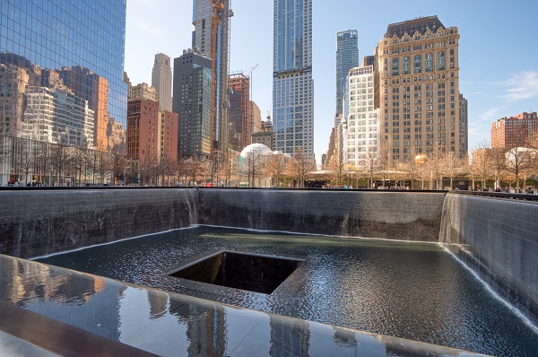 A view over the south pool of the 9/11 Memorial on a sunny day shows buildings reflecting in the water of the pool.