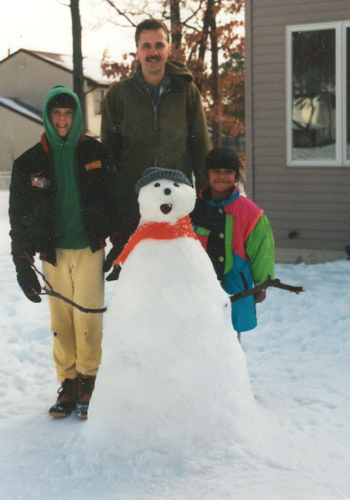 Michael Diehl stands in the snow next to a snowman with his two children, a boy and a girl. 