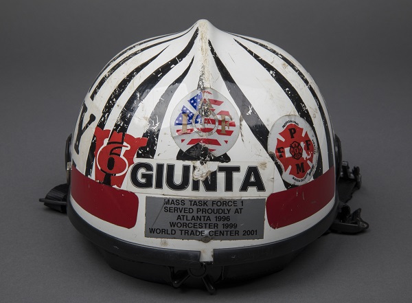A hard hat of rescue and recovery worker Gerry Giunta is displayed on a gray surface. The hard hat is zebra striped and says “Mass Task Force 1 served proudly at Atlanta 1996, Worcester 1999, and World Trade Center 2001.