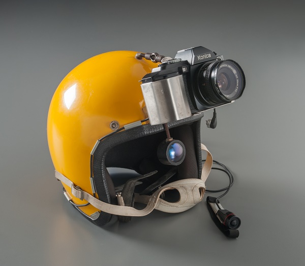 The yellow skydiving helmet of Charles W. Magee is displayed on a gray surface at the Museum. The helmet has a camera attached to the top of it and a white chin strap below.
