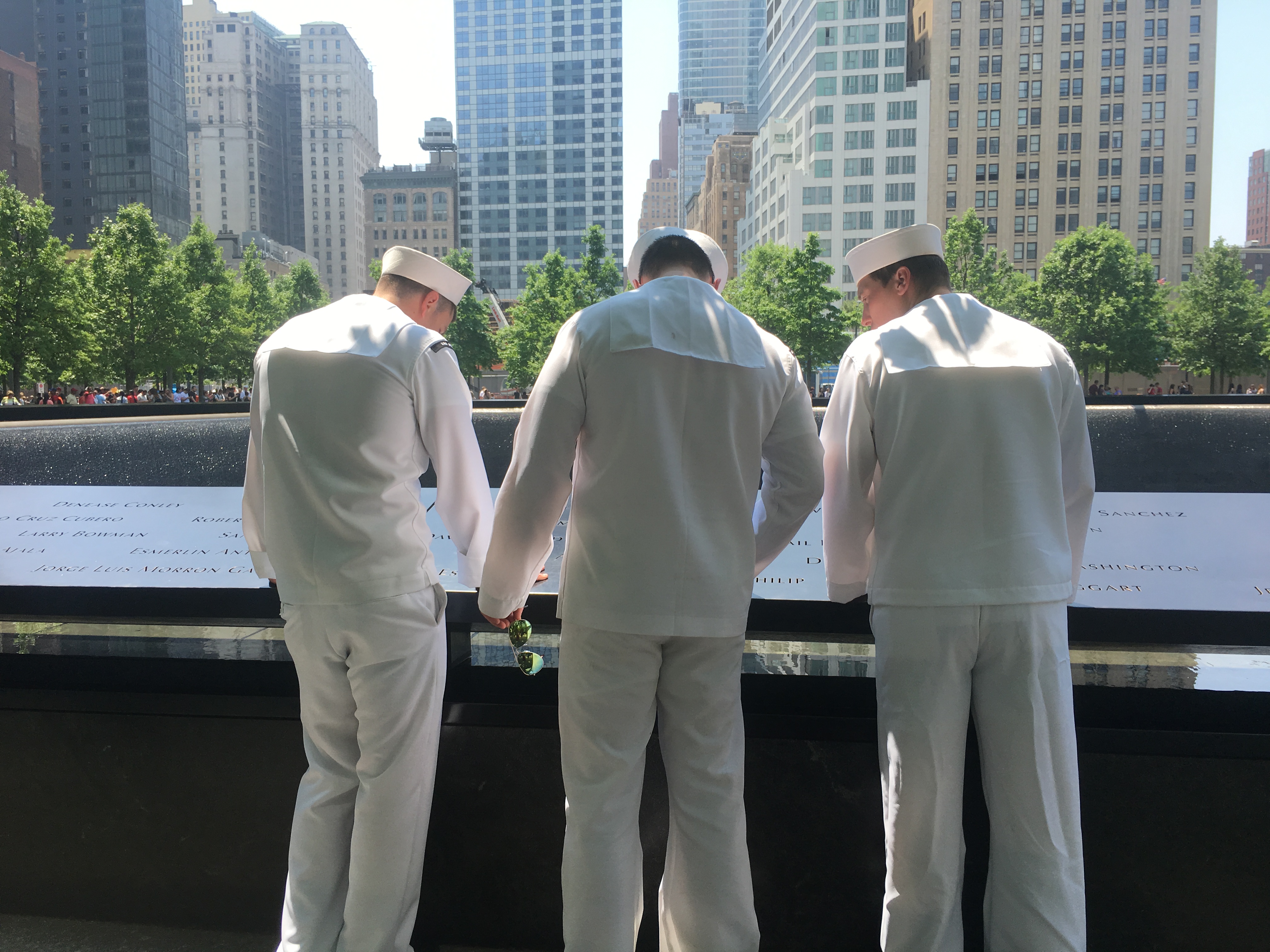 Three men in white U.S. Navy outfits look at a group of names on the 9/11 Memorial. They are turned away from the camera as they gather around the names on a bronze parapet.