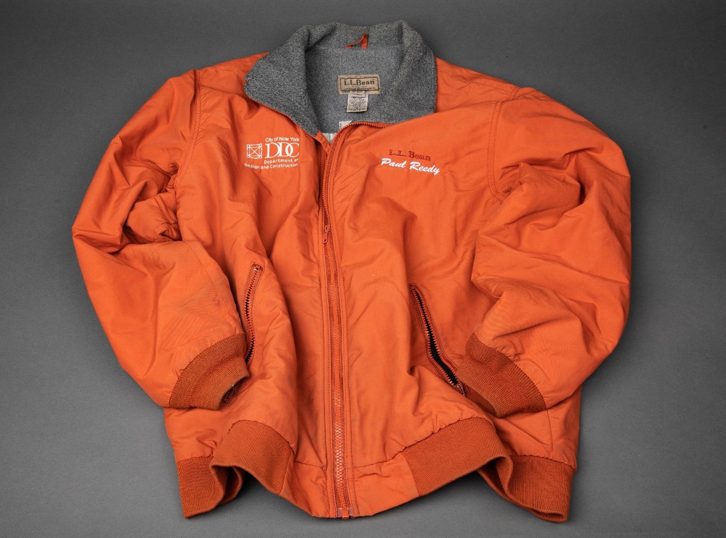 An orange L. L. Bean jacket work by Paul W. Reedy Sr. during his time at the New York City Department of Design and Construction is displayed on a gray surface. Reedy was in a leadership role at Ground Zero.