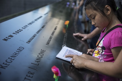 A girl stands at a bronze parapate etched with the names of victims. A woman’s arm is seen pointing to a piece of paper the girl is reading.