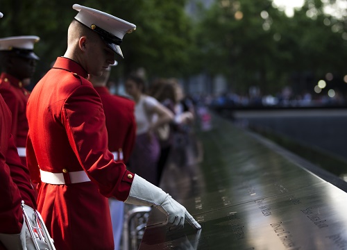 A Marine in a formal red outfit traces a name on the 9/11 Memorial with his gloved finger. Several other Marines stand behind him.