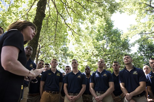 A woman speaks to a group of U.S. Marines under trees on the Memorial plaza.