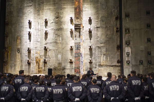 NYPD recruits stand in front of the slurry wall during a ceremony in the 9/11 Memorial Museum’s Foundation Hall. The recruits are facing away from the camera as the watch a speaker onstage. Their jackets read NYPD in large letters.