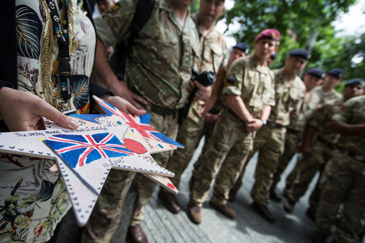 British Army Rugby Union players in camouflage stand together on Memorial plaza. One of them holds stars of hope that include drawings and messages. A star of hope in the foreground has a drawing of the British flag along with the message.