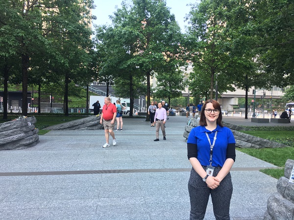 9/11 Memorial & Museum interpretive guide Katie Curran stands in front of the newly opened Memorial Glade. She’s wearing a blue shirt and an identification badge.