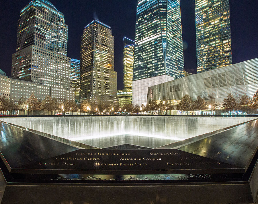 A reflecting pool of the 9/11 Memorial is seen illuminated at night. Surrounding buildings, including One World Trade Center, are lit up in the background.