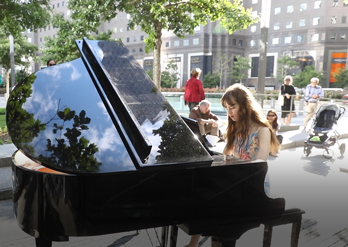 A young woman plays at a black grand piano on Memorial plaza as part of a special music program. Several visitors watch from behind her.