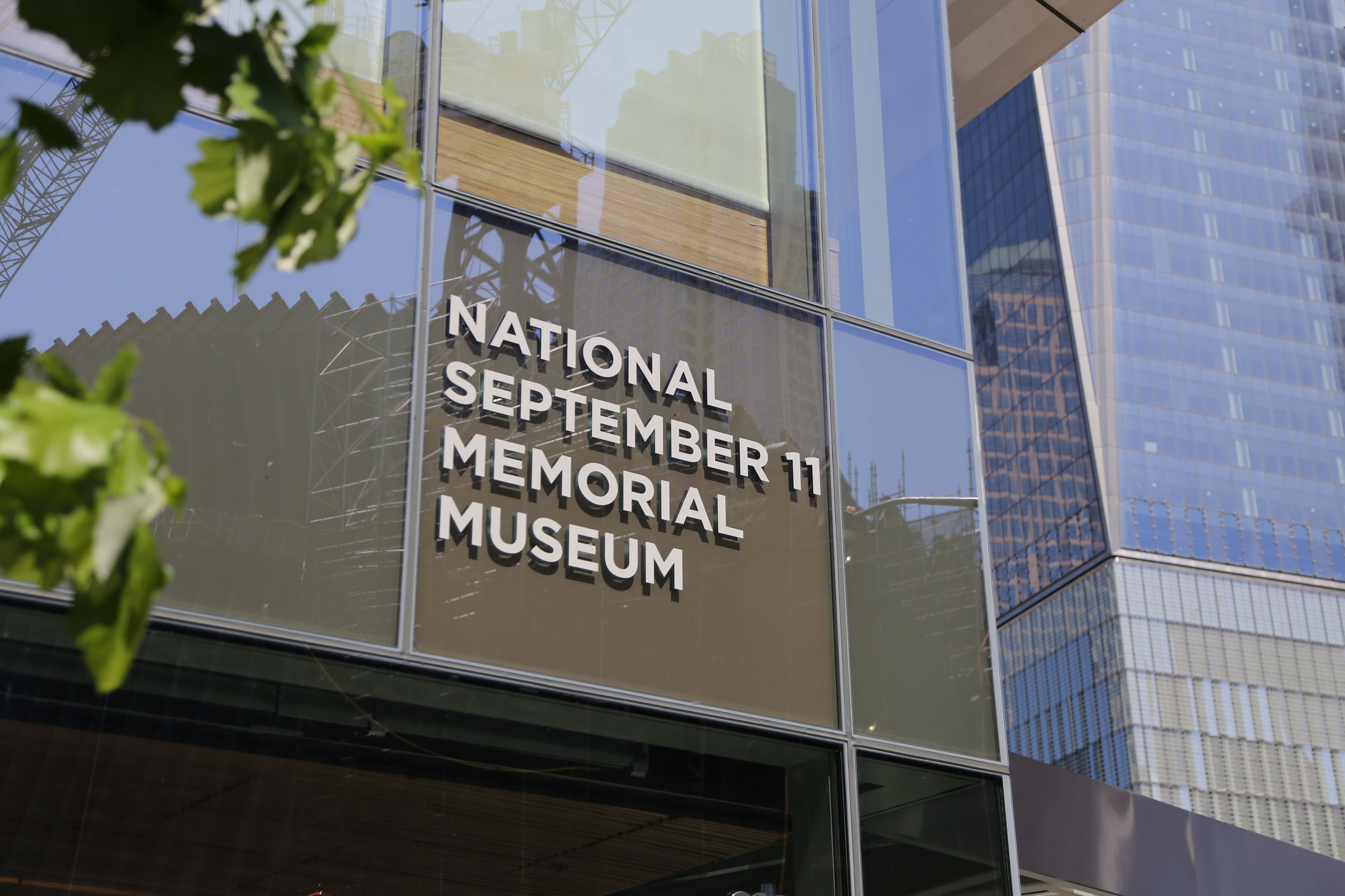 The entrance sign of the 9/11 Memorial Museum is displayed on the Museum’s glass facade. It reads “National September 11 Memorial Museum. One World Trade Center is visible in the background.