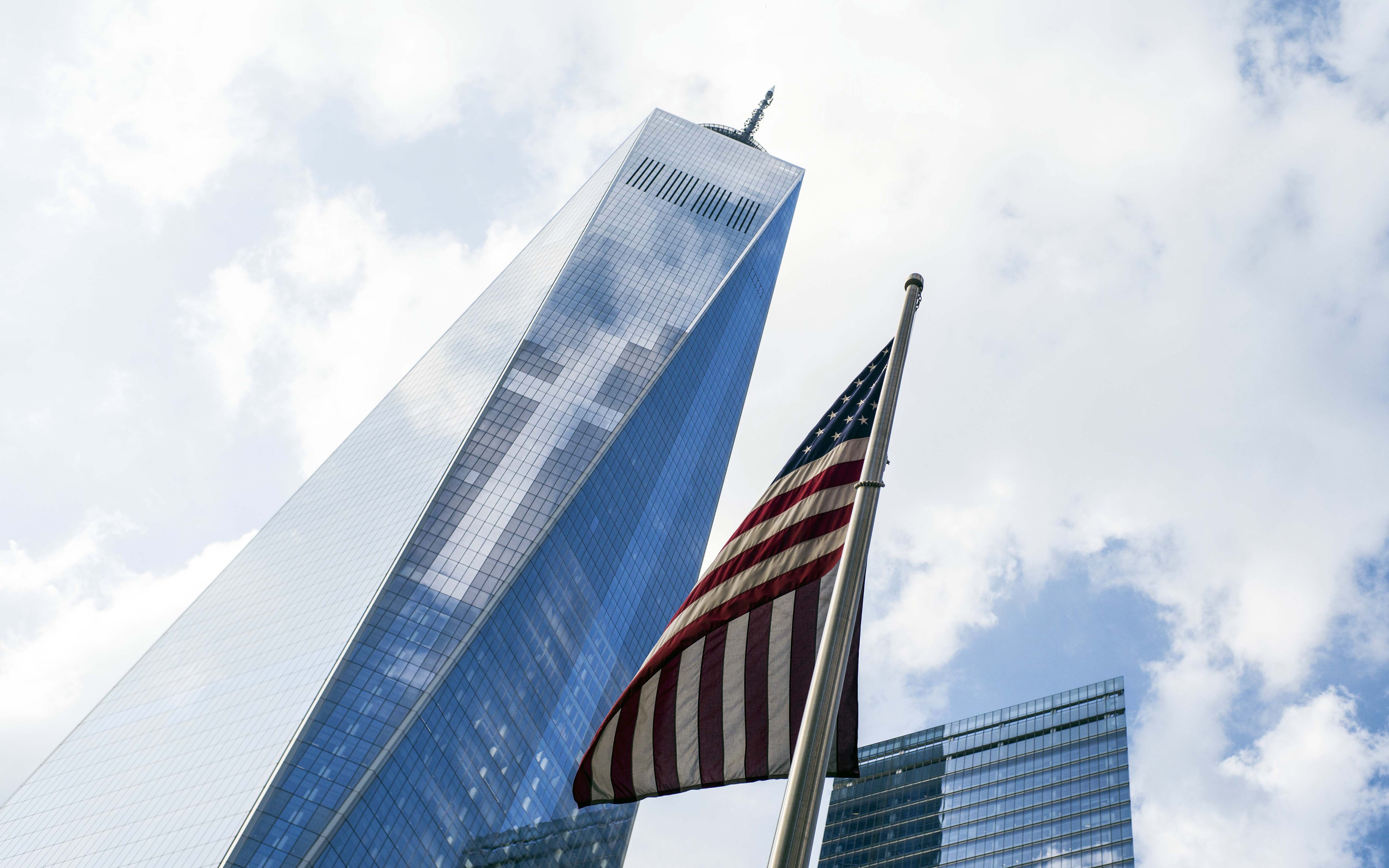 An American flag flies over the 9/11 Memorial in this image looking up at One World Trade Center on a cloudy day. The clouds are seen reflecting off the glass of One World Trade Center.