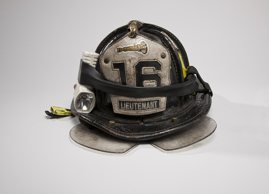 FDNY Lieutenant Mickey Kross’ helmet is displayed on a white surface at the Museum. It is equipped with a flashlight and a visor. The number 16 is on the front of it.