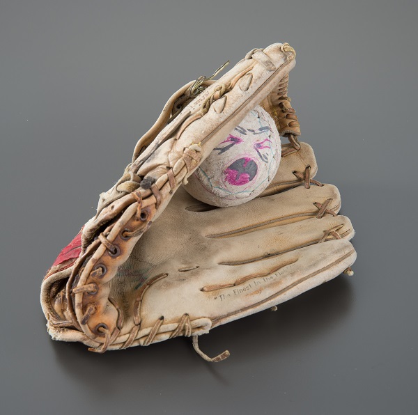 A softball and Rawlings brand glove belonging to FDNY member Robert Francis Wallace is presented on a gray surface. The softball has a screaming face drawn on it in black and pink marker.