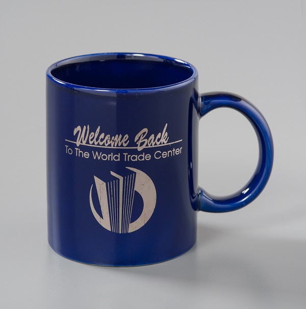 A blue “Welcome Back to the World Trade Center” mug is displayed on a white surface at the Museum. It includes an insignia of the Twin Towers.