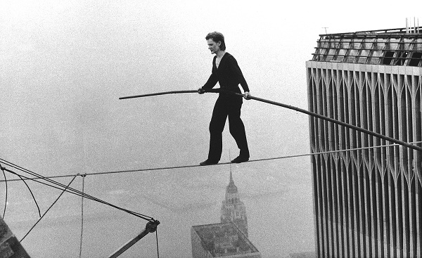 In this black-and-white historical photo, Philippe Petit holds a long pole as he does a hire-wire walk between the Twin Towers. The buildings of lower Manhattan can be seen below him.