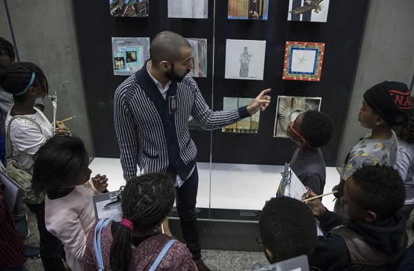 Middle school students from Macademy School of Science and Technology take part in a guided student program. An education specialist points out objects displayed in a glass case as the students look on.