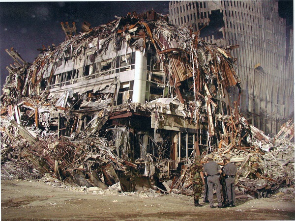 Several men in military and law enforcement garbs stand beside the smouldering remains of the Marriott World Trade Center hotel on a night shortly after the attacks. The hotel is covered in twisted debris from the Twin Towers.
