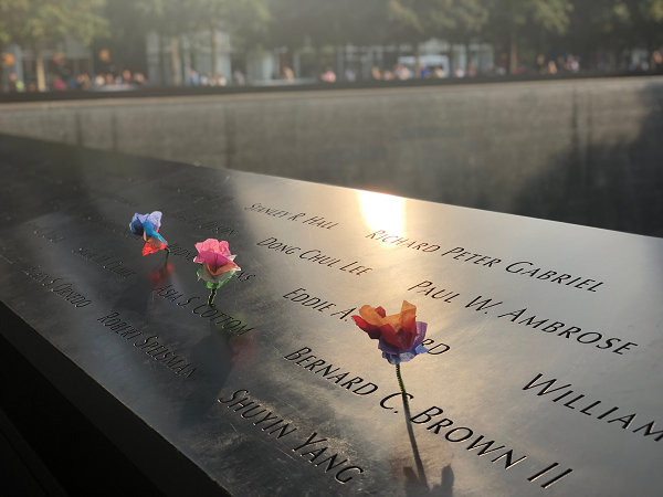 Several origami flowers have been placed at names on the 9/11 Memorial. A rising sun reflects off the bronze parapet where the names are etched.