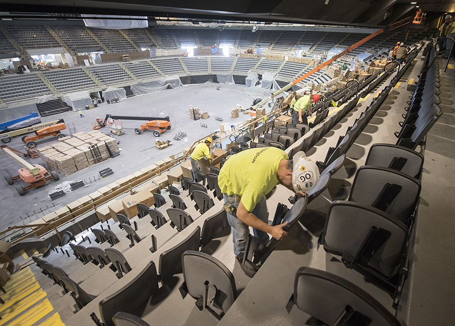 A man in a yellow shirt and white hardhat install seats at the newly renovated Nassau Coliseum on Long Island. Other workers install seats behind him. Construction machines are seen on the stadium floor in the background.