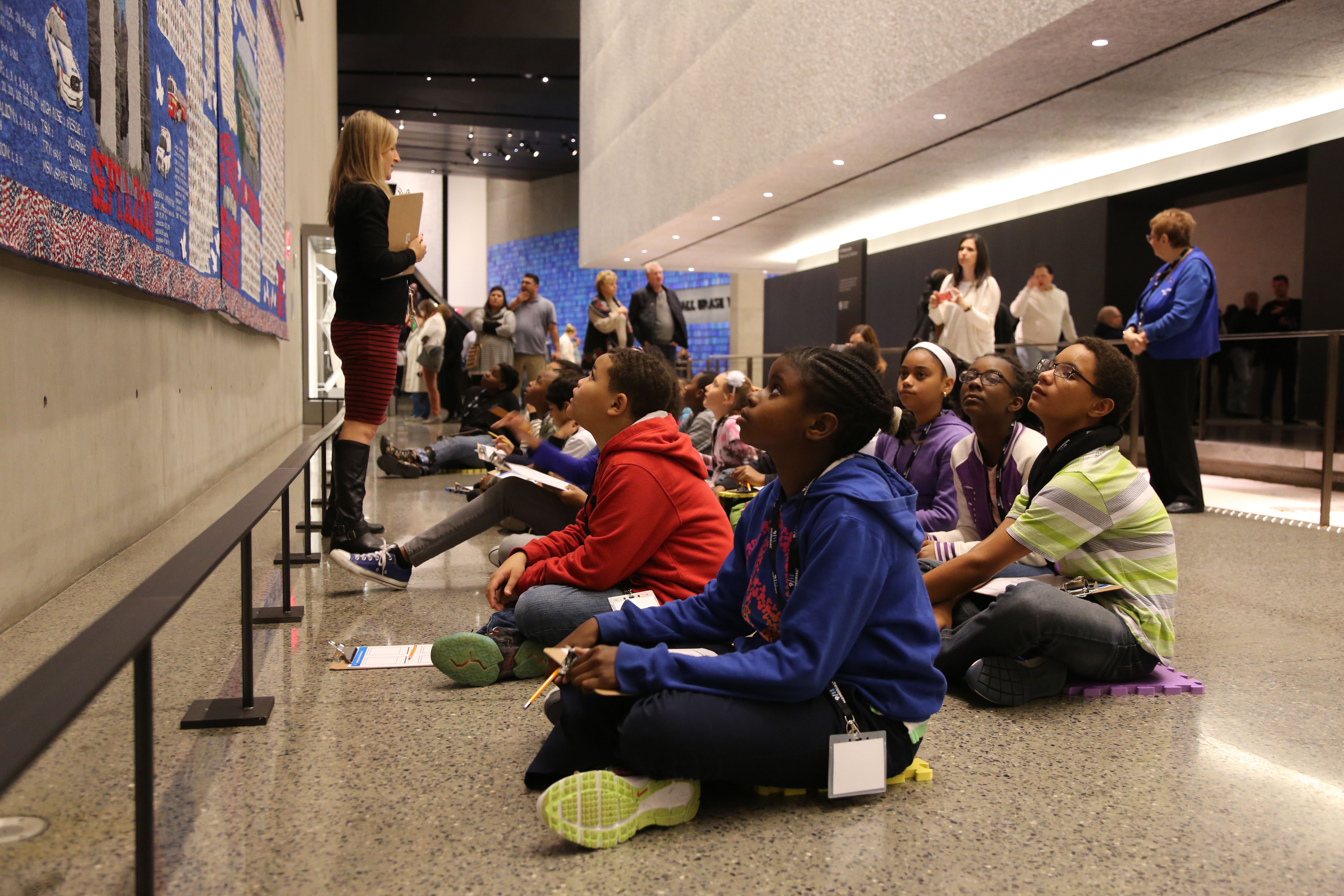 Students sit on the floor of the Museum as they listen to a woman standing in front of the Victims’ Memorial Quilt speak.