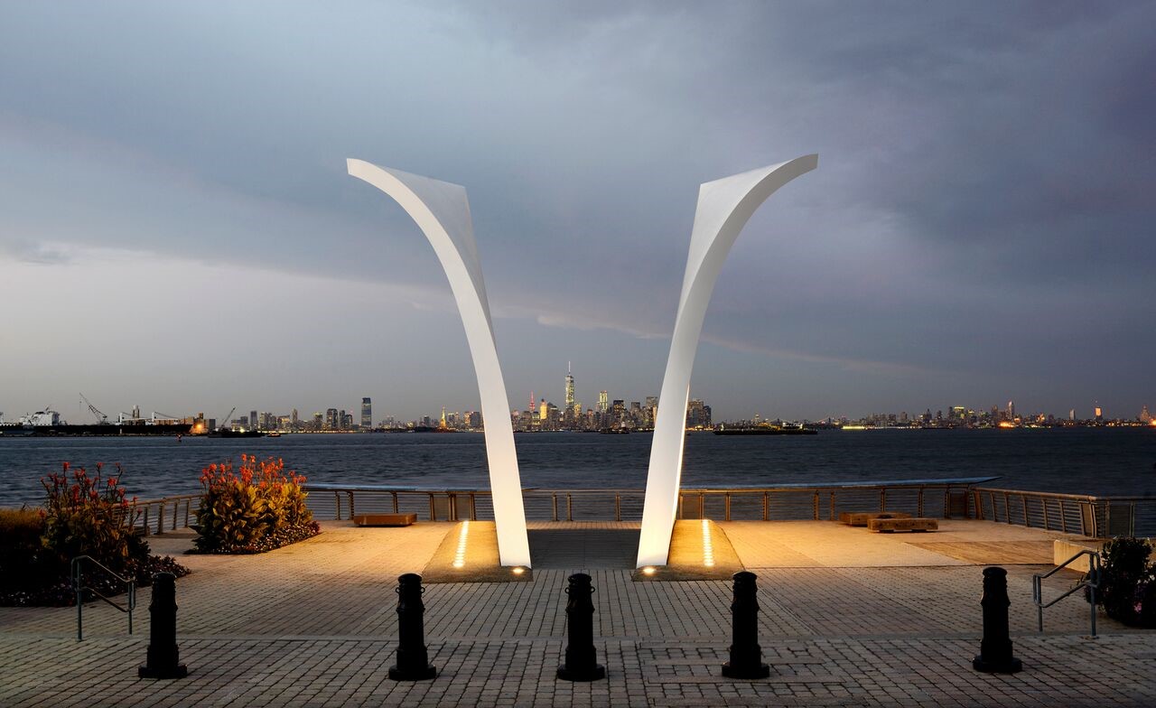 A memorial called “Postcards” is lit up on Staten Island at dusk. The memorial features two curved, white walls with tributes to the 263 Staten Island residents killed on 9/11 and the 1993 bombing of the World Trade Center. The skyline of Manhattan can be seen in the space between the two walls. The skyline of Jersey City is to the left, and the skyline of Brooklyn is to the right.
