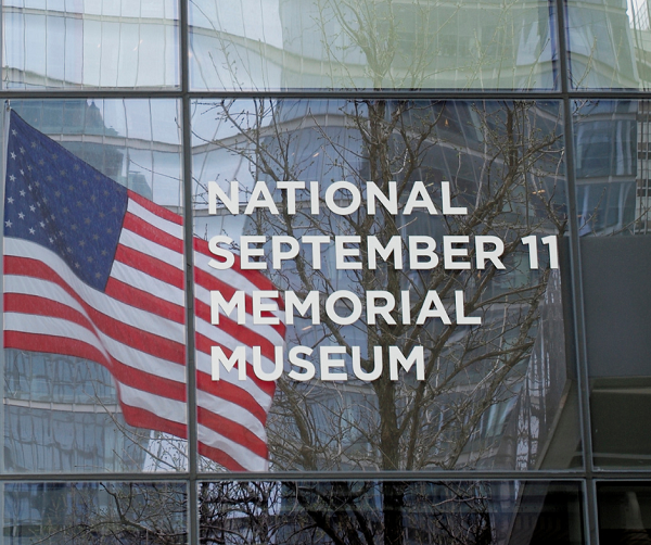 A sign on the Museum reads “National September 11 Memorial Museum.” An American flag reflects off the glass that the sign is displayed on.