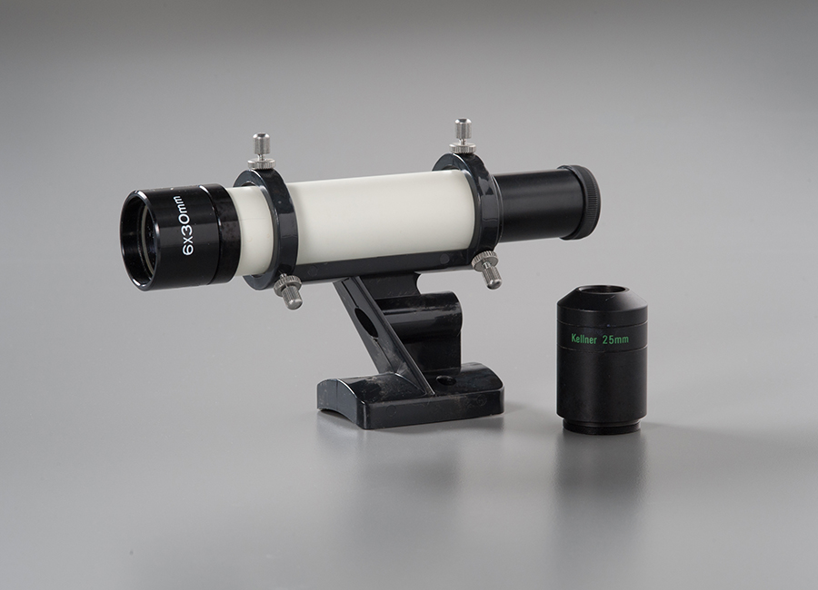 Howard Kestenbaum’s white and black telescope is displayed on a gray surface at the Museum. A lens sits beside the telescope.