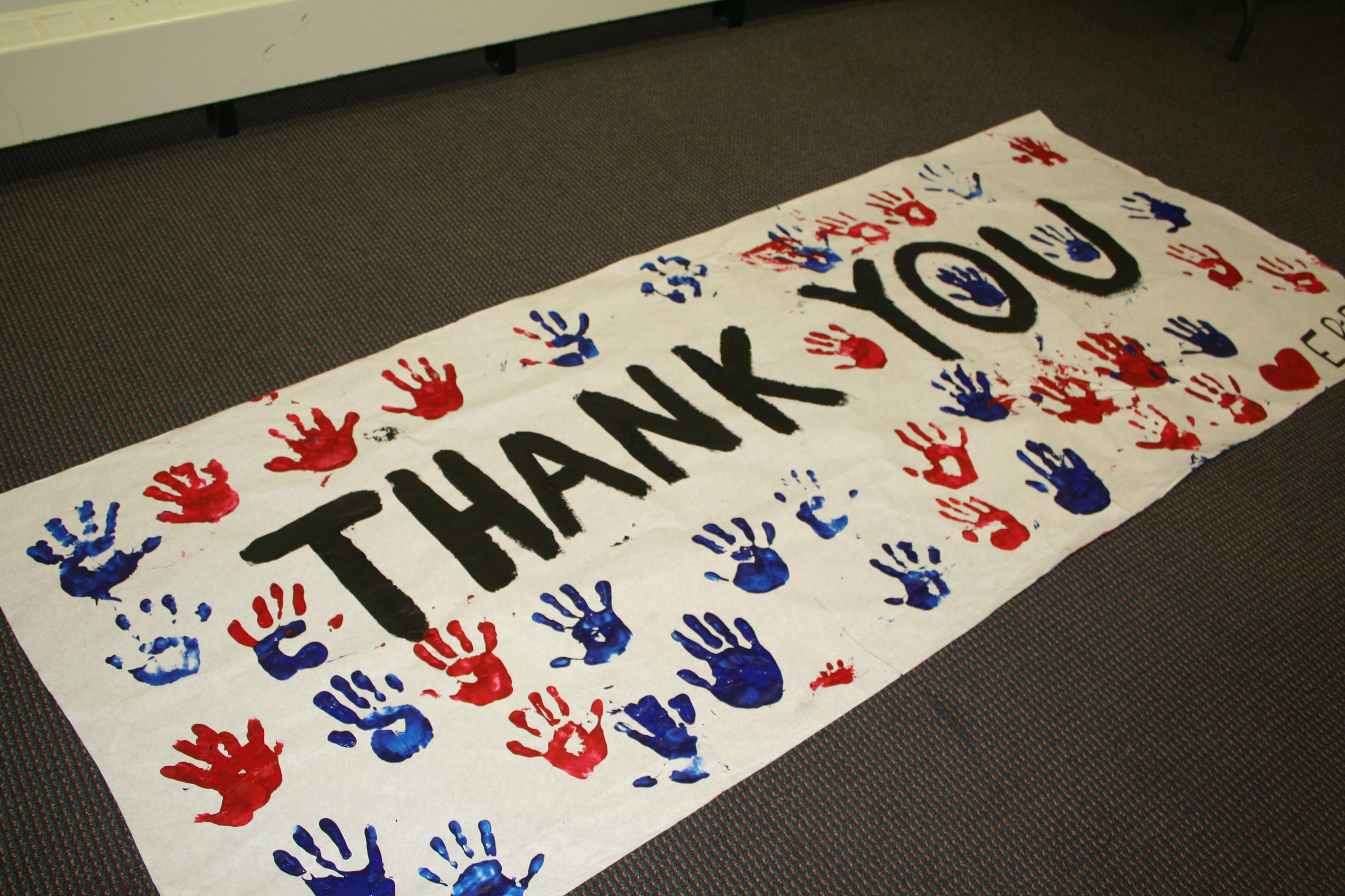 A “Thank You” poster for U.S. troops is displayed on a gray carpet. The poster includes red and blue handprints and the words “Thank You” hand painted in large letters.