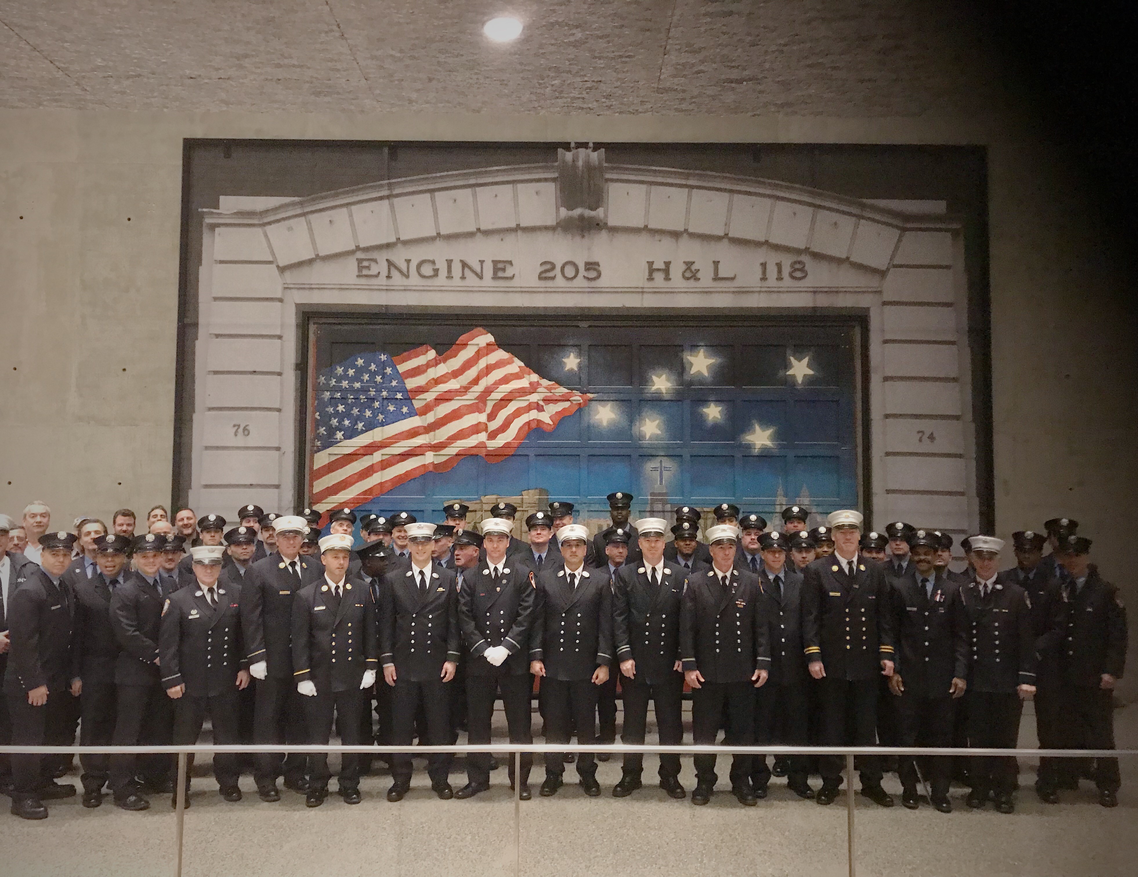 At the Museum’s Tribute Walk, members of FDNY Engine 205 and Ladder 118 stand in front of a firehouse door painted in tribute to their fellow firefighters killed on 9/11. The painting depicts an American flag on a starry night.