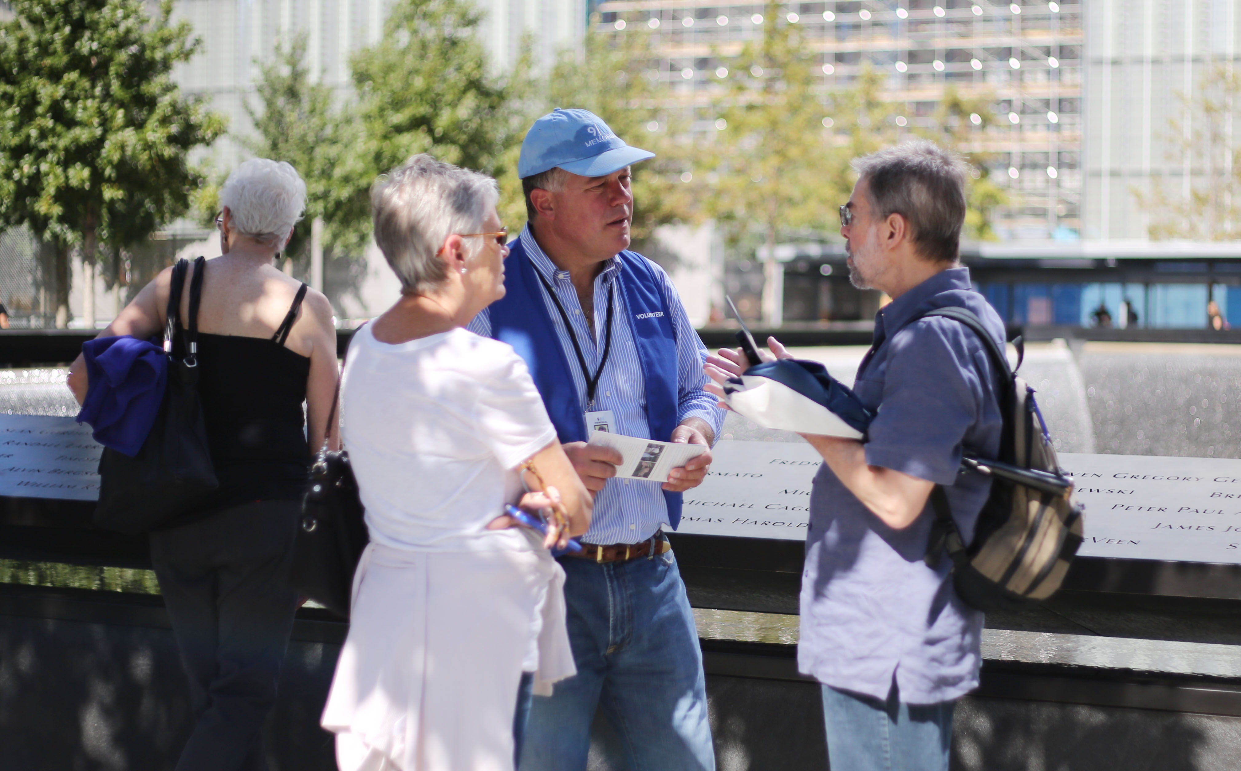 A volunteer speaks with two visitors beside a reflecting pool on the 9/11 Memorial plaza.