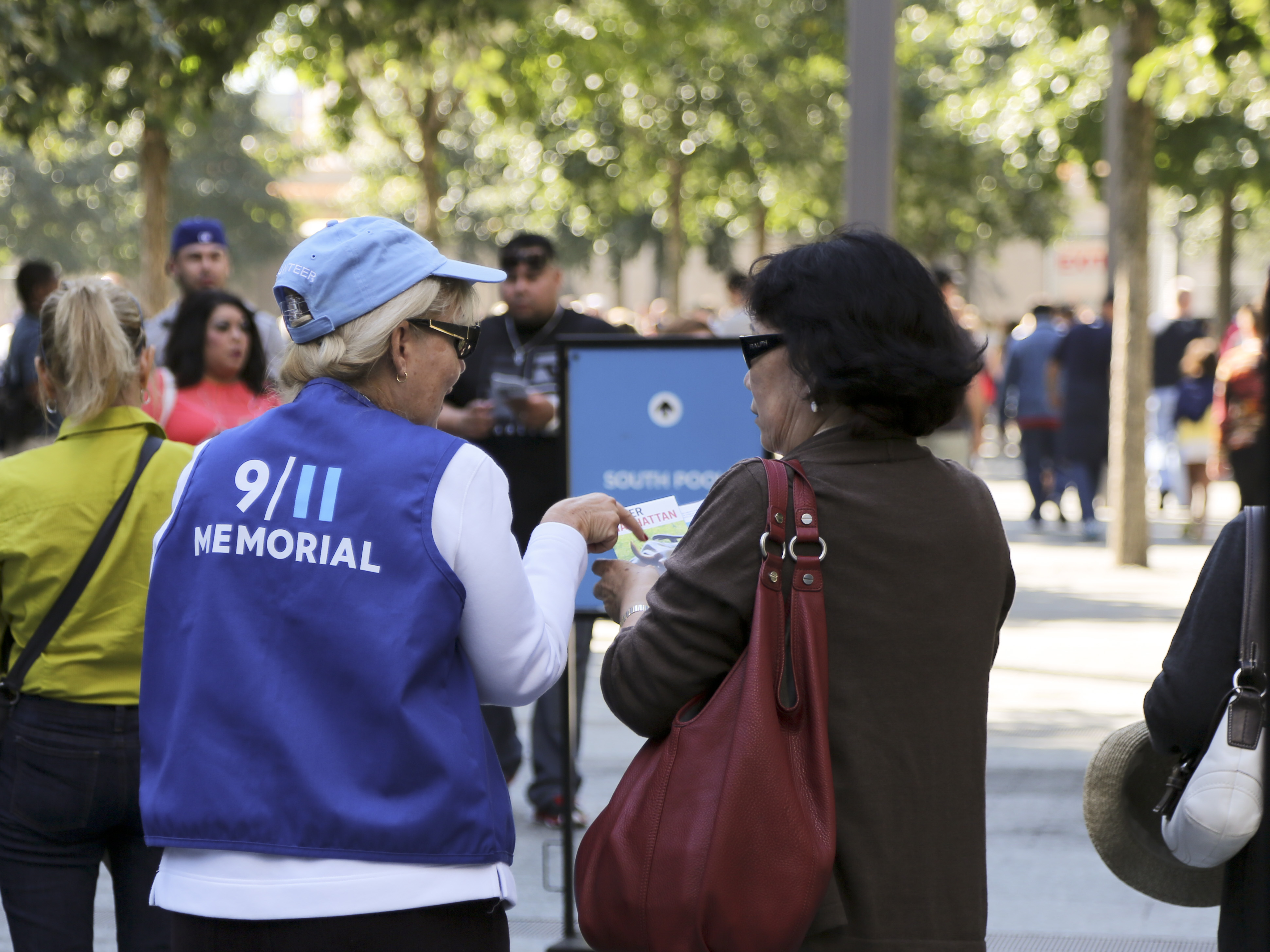 A volunteer in a blue vest that reads 9/11 Memorial assists a woman on the Memorial plaza.