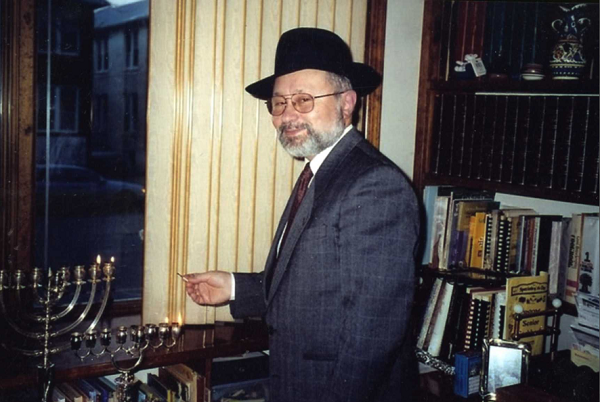 Abraham Zelmanowitz lights a menorah during the Hanukah of 2000. He is wearing a suit and tie and a hat and standing next to a window.
